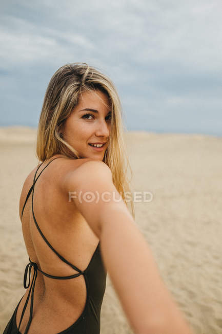 Jovem alegre em maiô sorrindo e olhando para a câmera enquanto lidera caminho na praia de areia — Fotografia de Stock