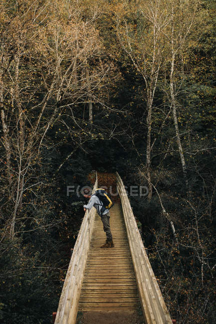Young guy with backpack leaning on railing of ancient bridge near autumn forest — Stock Photo