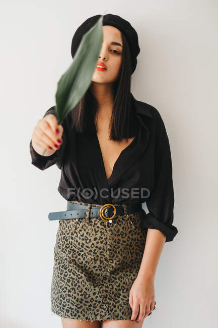 Jovem fêmea encantadora em roupa elegante olhando para a câmera e cobrindo o peito com folha de planta verde, enquanto está perto da parede branca — Fotografia de Stock