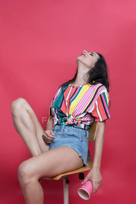 Passionnée jeune femme en chemise rayée colorée posant sur chaise avec tasse en papier — Photo de stock