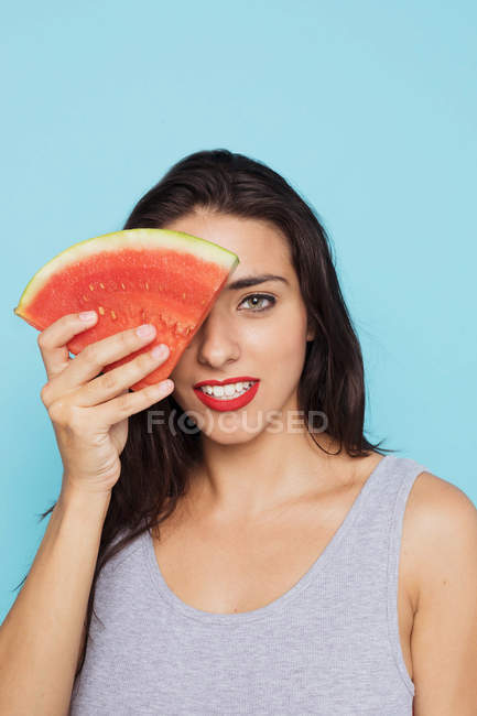 Young woman covering eye with piece of watermelon on blue background — Stock Photo