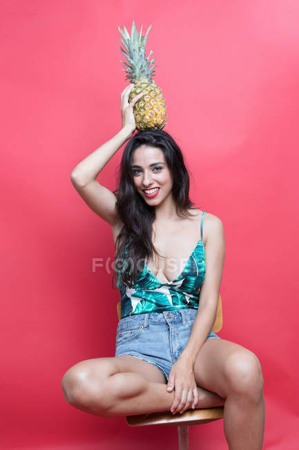 Young woman posing on chair with pineapple on head on pink background — Stock Photo