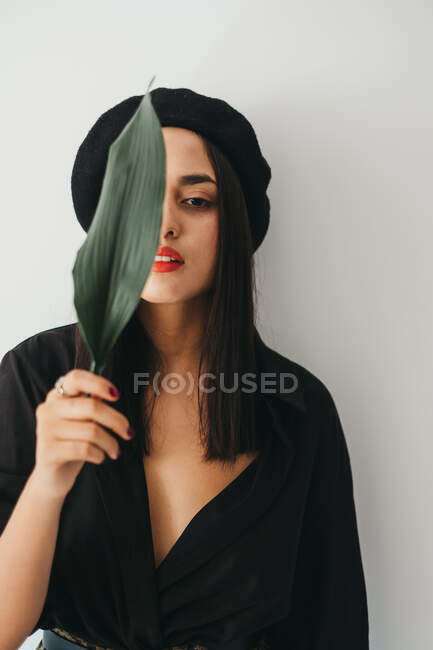 Очаровательная молодая женщина в стильном наряде смотрит в камеру и покрывает грудь зеленым листком растения, стоя возле белой стены — стоковое фото