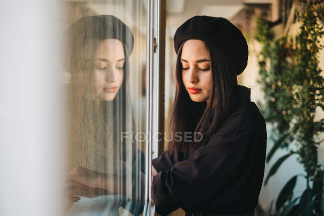 Seitenansicht der schönen jungen Frau in stilvollem Outfit am Fenster in gemütlichem Raum stehen und sich ausruhen — Stockfoto