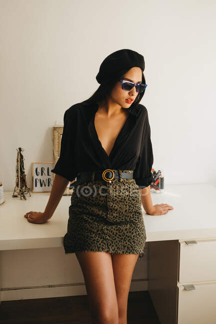 Attractive young woman in stylish outfit looking at camera while leaning on desk in cozy room — Stock Photo