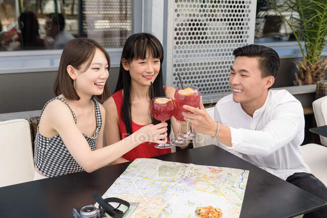 Três asiáticos sorrindo e batendo copos de bebida deliciosa enquanto sentado à mesa da câmera perto do mapa — Fotografia de Stock