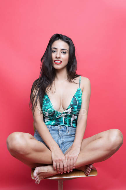 Portrait of young woman in patterned top and denim shorts sitting on chair on pink background — Stock Photo