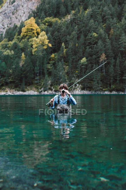 Jeune homme avec canne à pêche debout dans l'eau calme du lac pittoresque et boire — Photo de stock