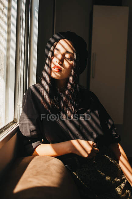 Чарівна молода леді в стильному вбранні тримає очі закритими і розслаблюючими, спираючись на диван біля вікна — стокове фото
