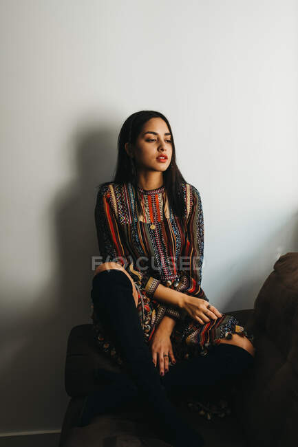 Schöne junge Brünette in stilvollem Outfit stützt den Kopf und hält die Augen geschlossen, während sie auf einer bequemen Couch in der Nähe der weißen Wand sitzt — Stockfoto