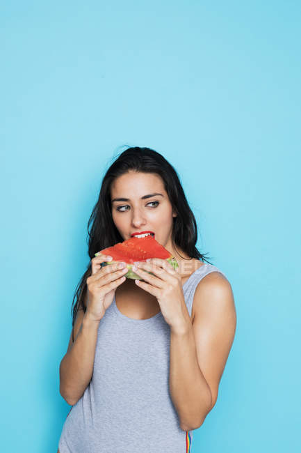 Sensuelle femme brune en body gris mangeant pastèque fraîche et regardant loin sur fond bleu — Photo de stock