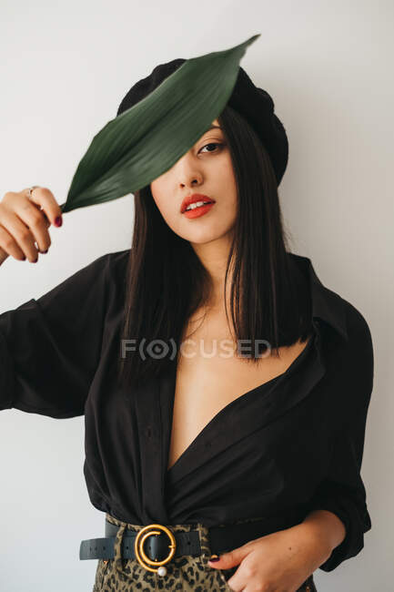 Чарівна молода жінка в стильному вбранні дивиться на камеру і покриває груди зеленим листям рослини, стоячи біля білої стіни — стокове фото