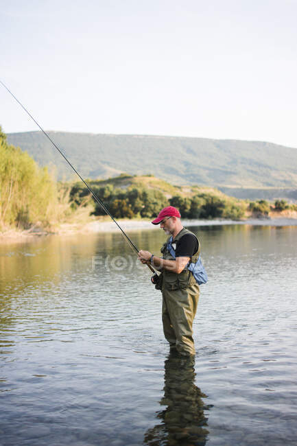 Vista laterale dell'uomo in trampolieri e cap pesca in acque tranquille con paesaggio pittoresco — Foto stock