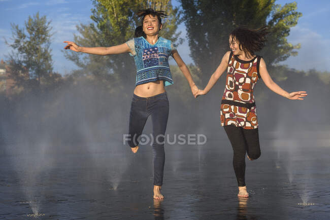 Two Asian ladies laughing and holding hands while running on water of splashing fountain in park — Stock Photo
