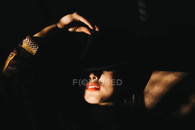 Attractive young female covering half of face with hat while standing in dark room — Stock Photo