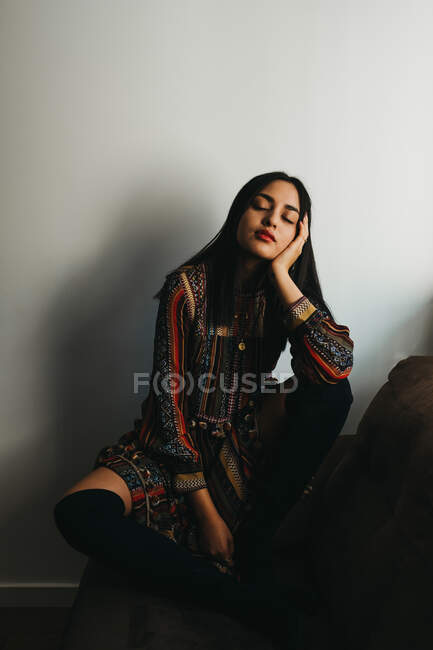 Bella giovane bruna in abito elegante che sostiene la testa e tiene gli occhi chiusi mentre si siede su un comodo divano vicino alla parete bianca — Foto stock