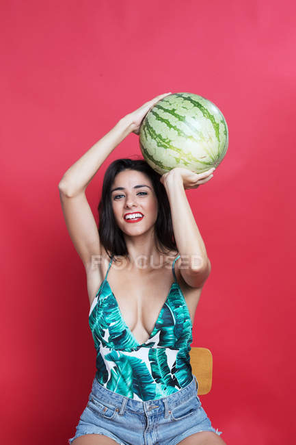 Jovem mulher em calções de ganga e top segurando melancia acima da cabeça — Fotografia de Stock