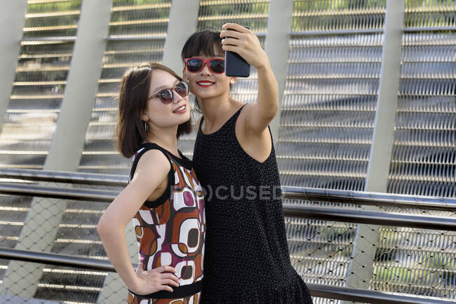 Duas encantadoras senhoras asiáticas sorrindo e posando para selfie enquanto estava perto de cerca de metal no parque moderno — Fotografia de Stock