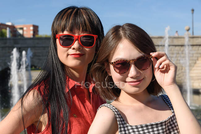 Dos preciosas damas asiáticas con gafas de sol elegantes sonriendo y mirando a la cámara mientras están de pie cerca de la fuente en el día soleado en el parque - foto de stock