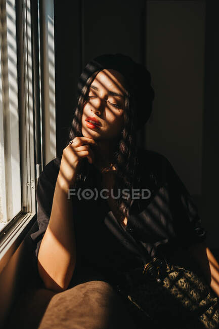 Чарівна молода леді в стильному вбранні тримає очі закритими і розслаблюючими, спираючись на диван біля вікна — стокове фото