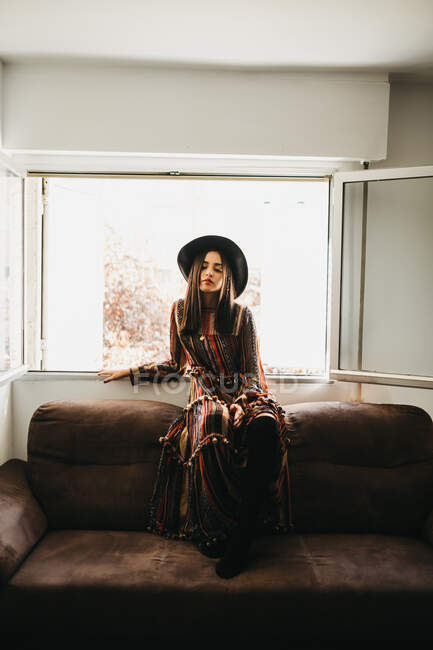 Attraktive junge Dame in stylischem Outfit lächelt und hält die Augen geschlossen, während sie auf der Rückseite des weichen Sofas am Fenster sitzt — Stockfoto