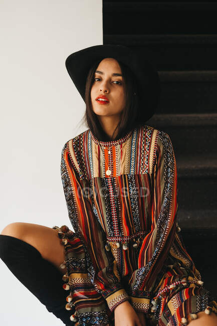 Beautiful young lady in stylish hat and dress supporting head and looking at camera while sitting on stairs at home — Stock Photo
