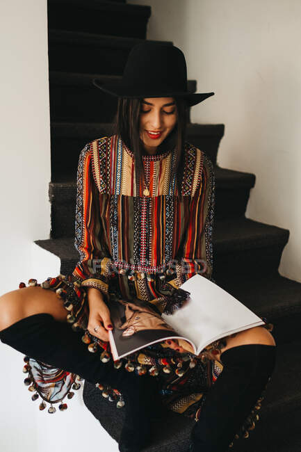 Atractiva joven hembra en sombrero elegante y vestido mirando a la cámara mientras se sienta en los pasos y la lectura de la revista de moda - foto de stock