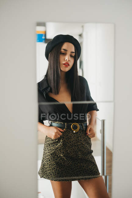 Jolie jeune femme en tenue élégante regardant miroir mural tout en se tenant dans une chambre confortable à la maison — Photo de stock