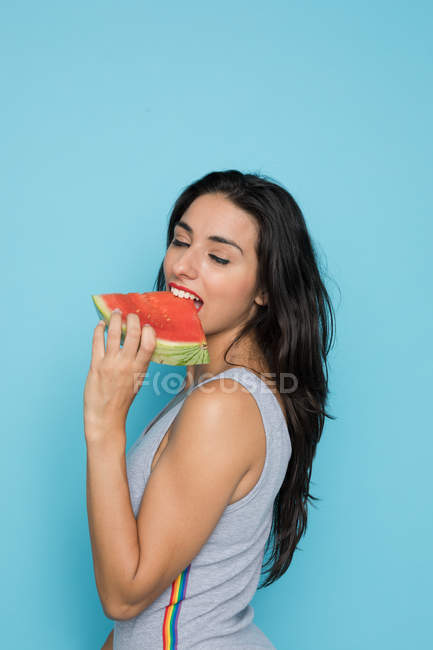 Sensual morena mulher em cinza bodysuit comer melancia fresca e olhando para o fundo azul — Fotografia de Stock