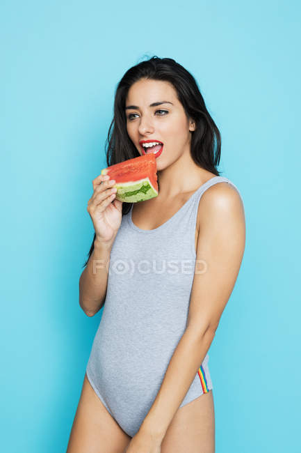 Sensuale donna bruna in body grigio mangiare anguria fresca e distogliere lo sguardo su sfondo blu — Foto stock