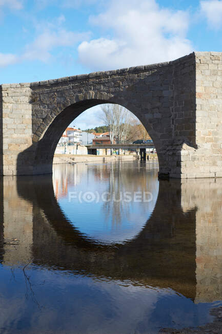 Arco del viejo puente de piedra con el río todavía tranquilo en el fondo del cielo azul nublado - foto de stock