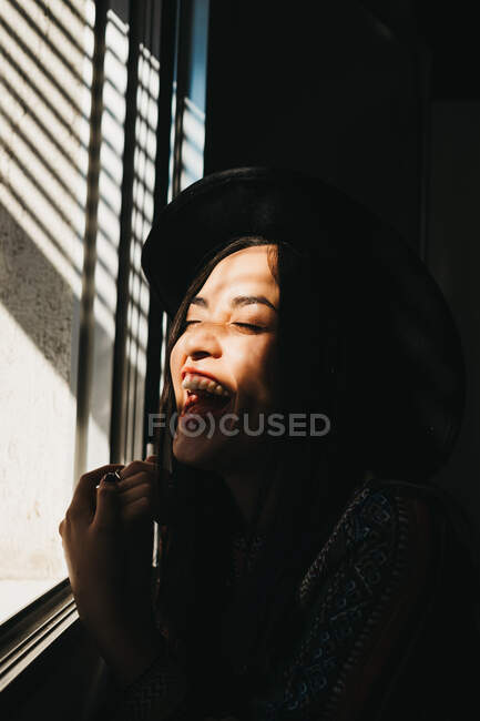 Lovely young female keeping eyes closed and supporting head while sitting near window in dark room and enjoying bright sunlight — Stock Photo