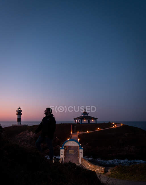 Handsome male looking away while standing on shore near illuminated bridge on background of beautiful islet with lighthouse and cloudless evening sky RELEASE — Stock Photo