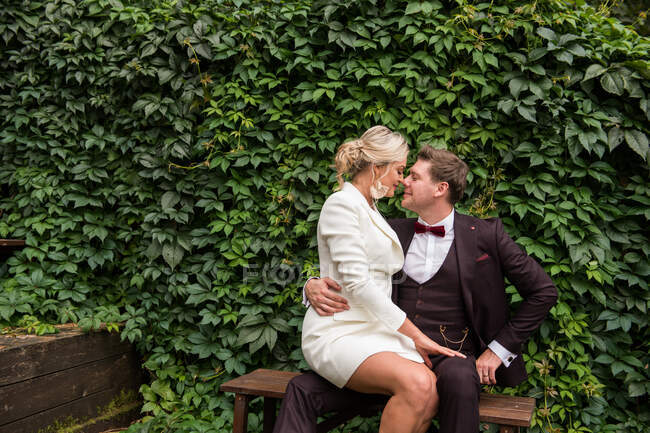 Excited man and woman wearing trendy wedding suits and looking at each other against green hedge — Stock Photo