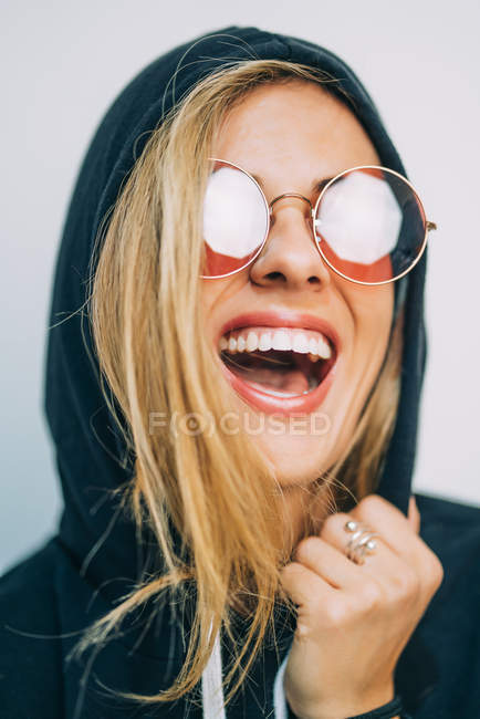 Jovem loira de óculos de sol e capuz rindo no fundo branco — Fotografia de Stock
