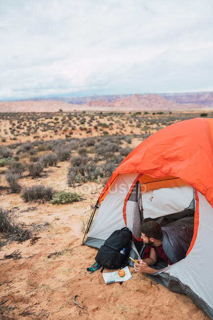 Crop uomo barbuto mangiare mela fresca e la navigazione smartphone moderno mentre sdraiato in tenda durante il campeggio nel deserto — Foto stock