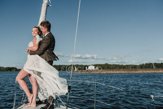 Vue latérale du couple marié heureux debout sur le nez du yacht et embrassant sur le lac — Photo de stock