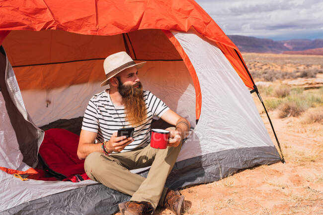 Бородатий хлопець у повсякденному вбранні тримає кухоль гарячого напою та сучасний смартфон, сидячи біля намету та дивлячись у чудову природу — стокове фото