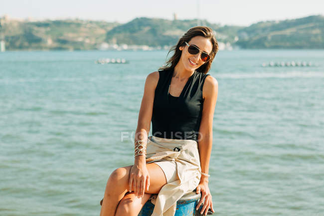 Mulher na moda sentado no toco à beira-mar com barcos à vela no fundo — Fotografia de Stock