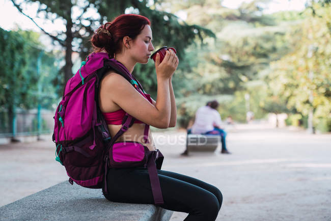 Young woman in sportswear with pink backpack sitting on bench in park and drinking hot tea from metal cup — Stock Photo