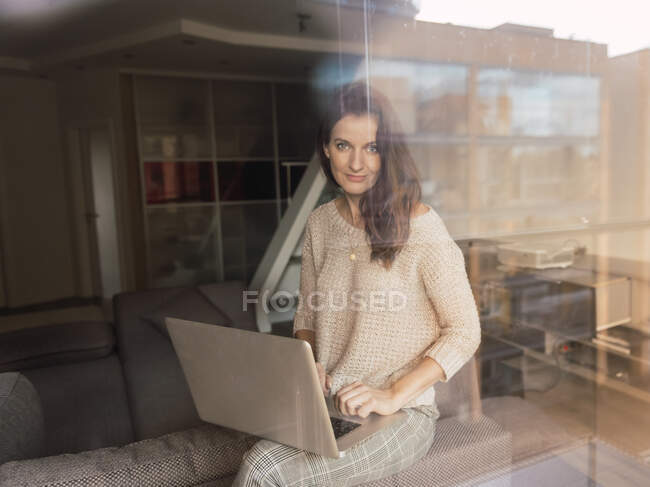 Hübsche erwachsene Frau in elegantem Outfit stöbert im modernen Laptop und schaut in die Kamera, während sie in einem gemütlichen Raum hinter riesigen Fensterscheiben sitzt — Stockfoto