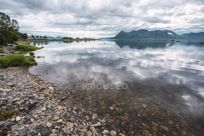 Paesaggio di calmo lago trasparente con costa ricoperta di piccole pietre sullo sfondo di montagne e cielo nuvoloso — Foto stock