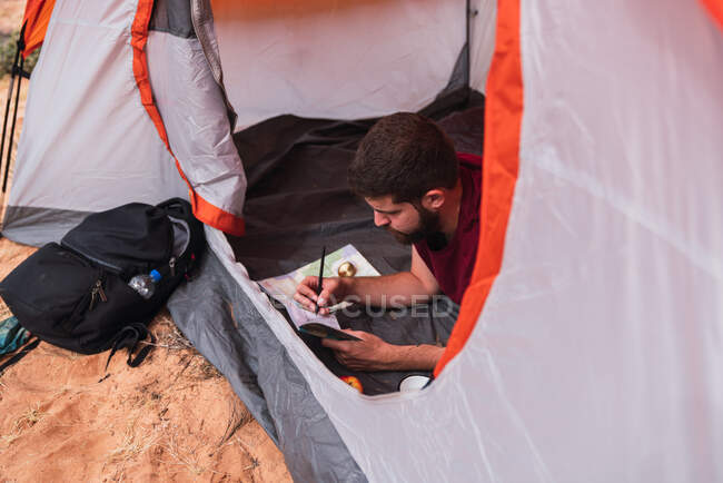 De cima tiro de cara jovem deitado na tenda e desenho em caderno de esboços enquanto acampando no deserto — Fotografia de Stock