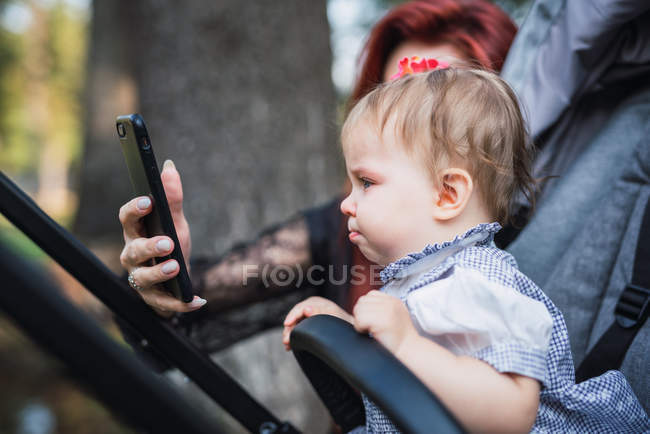 Невпізнана жінка тримає сучасний смартфон і намагається підбадьорити чарівну сумну дівчинку в колясці — стокове фото