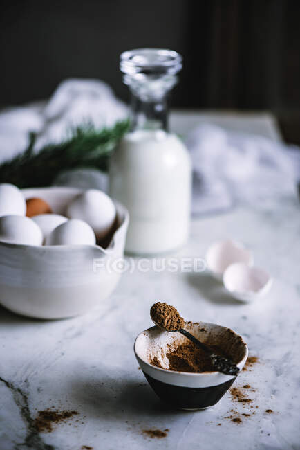 Bol et cuillère de cacao en poudre debout sur une table en marbre près d'un bol d'œufs frais et une bouteille de belle laiterie — Photo de stock