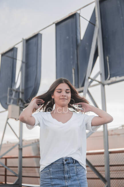 Чарівна молода жінка в білій сорочці і джинсовій спідниці погладжує волосся, що стоїть на міському фоні — стокове фото