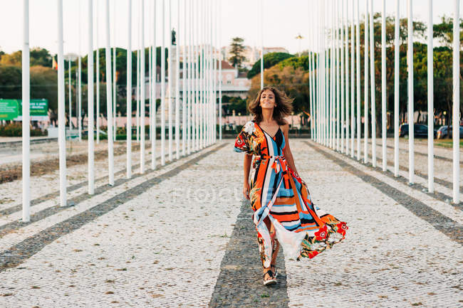 Mujer contenta en vestido ornamental colorido caminando en el paseo pavimentado sonriendo a la cámara - foto de stock