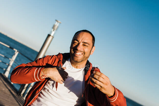 Похмурий дорослий самець усміхається і жестикулює, стоячи на пірсі біля моря в чудовий сонячний день. — стокове фото