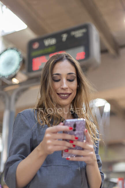 De dessous plan de jeune femme attrayante souriant et naviguant smartphone moderne tout en se tenant debout sur fond flou de la gare — Photo de stock