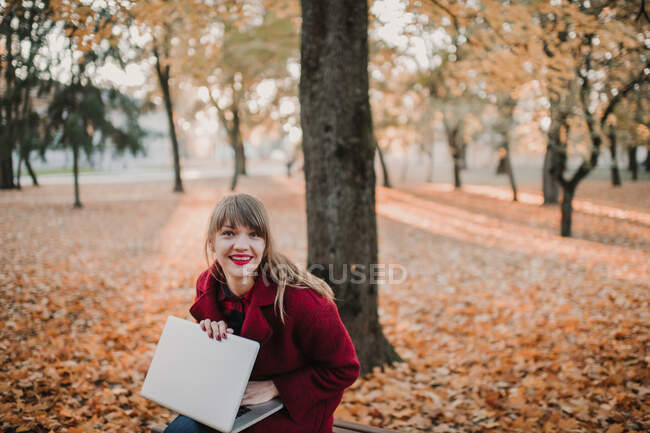 Jeune femme en manteau rouge à l'aide de l'appareil et assis sur le siège dans la forêt d'automne — Photo de stock
